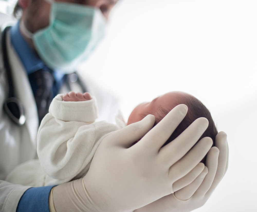 Male pediatrician holding a newborn baby while wearing mask and gloves.