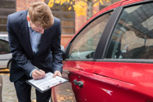 Insurance Agent Writing On Clipboard While Examining Car After Accident