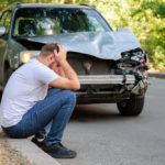 Man holding his head sitting in front of his car after a car accident.