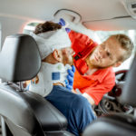 Medic applying bandage on the head of victim with serious damages sitting on the driver seat after the road accident. 