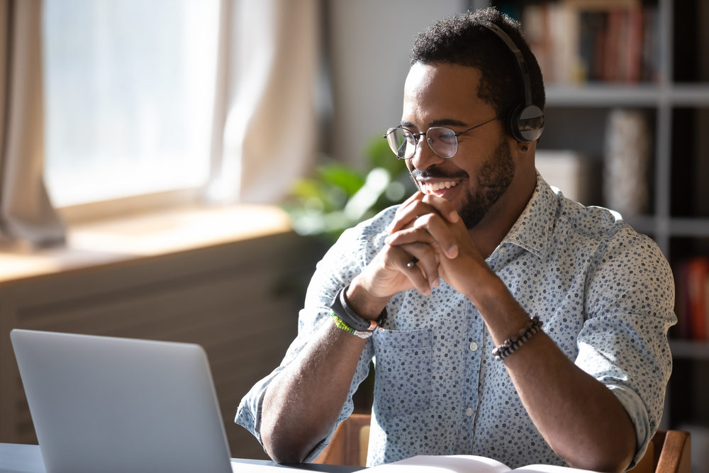 african american man in glasses wearing headphones watching a webinar on his laptop at a desk inside