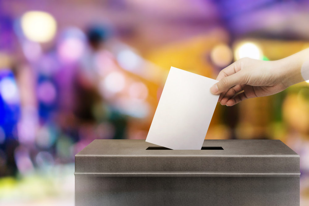 A hand drops a ballot into a large black box. Colorful blurred background.