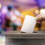 A hand drops a ballot into a large black box. Colorful blurred background.
