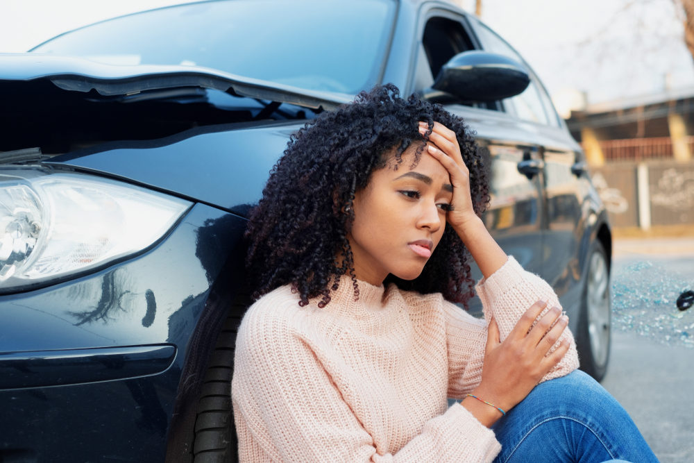 Black woman leaning against a car holding her head after a crash