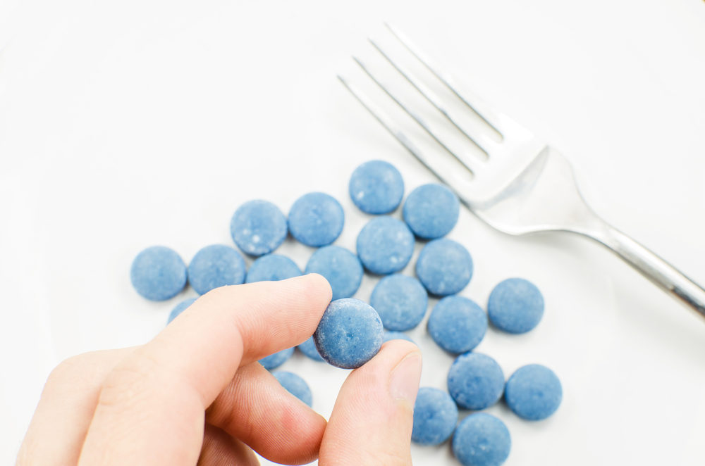 Blue Pill held in hand with fork and more on a plate in the background