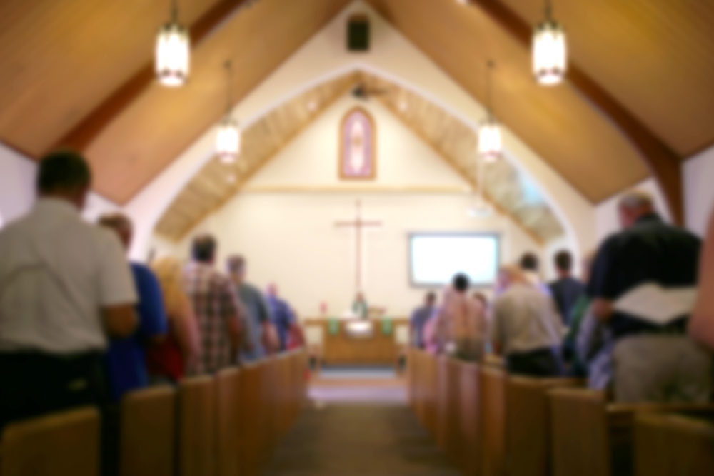 A blurred photo of the inside of a church sanctuary that is filled with people in the pews, and the pastor stands under a large cross at the altar.