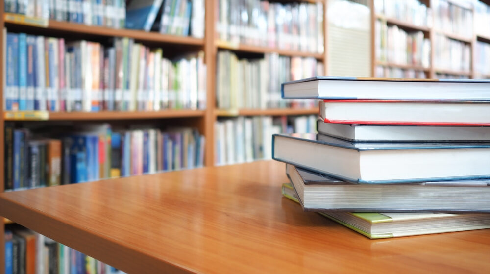 Delaware Brandywine School District Psychologist Arrested on Child Rape Charges Books stacked on wooden desk with blurred bookshelf in the background oflibrary