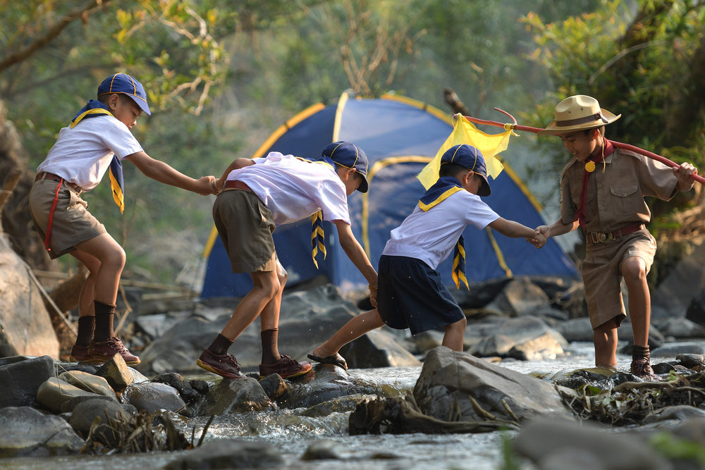 Young boy scouts help each other cross a river by stepping on rocks