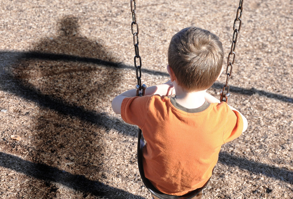 A young boy is sitting on a swing set and looking at a shadow figure of a man or bully at a playground