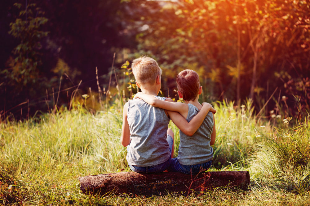 Two young boys with their arms around each other sit on a log