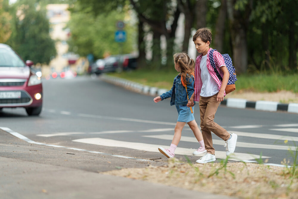 brother and sister holding hands walking through a crosswalk as a car approaches