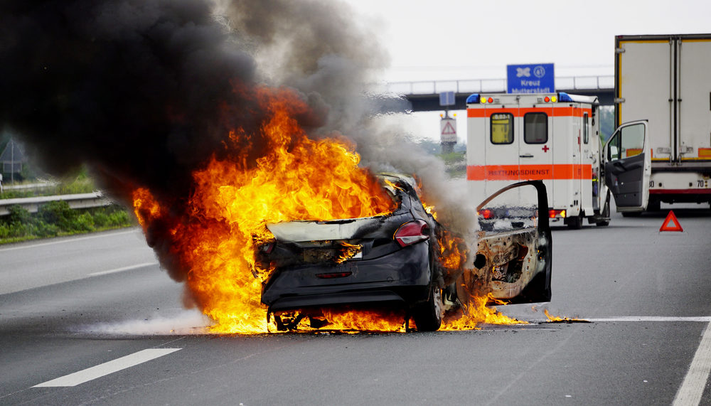 A car is ablaze on the highway with an ambulance in the background