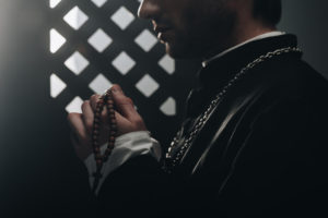 partial view of catholic priest holding wooden rosary beads near confessional grille in dark with rays of light
