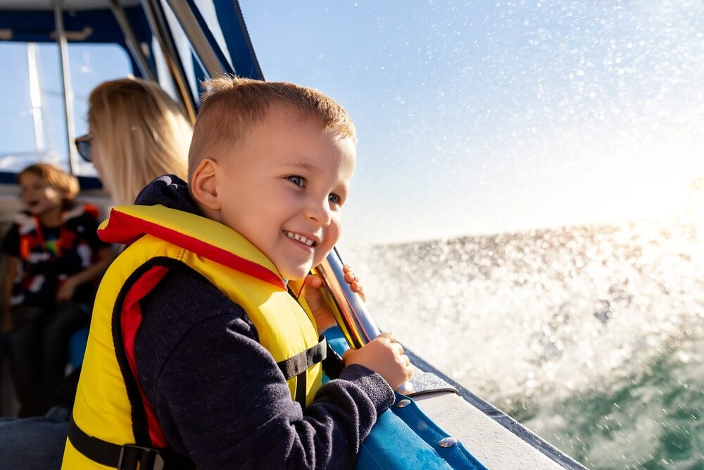 little boy in a life vest boating with his family as water splashes along side of the vessel