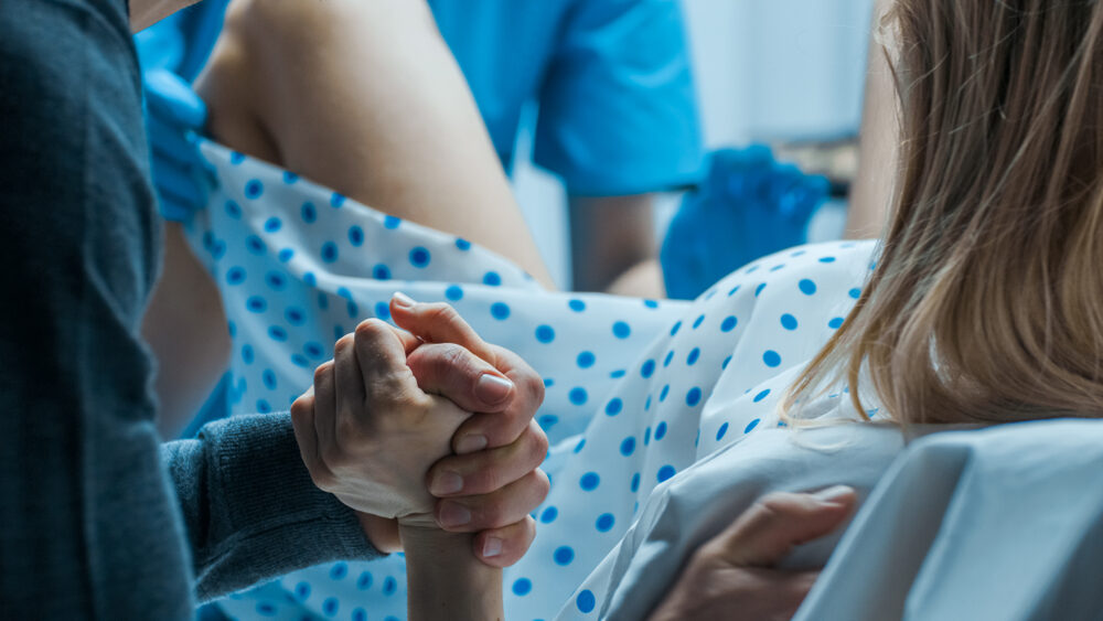 closeup of a woman is giving birth in hospital with husband holding her hand while doctor helps delivery