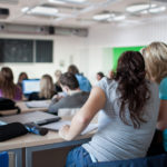 young pretty female college student sitting in a classroom full of students during class