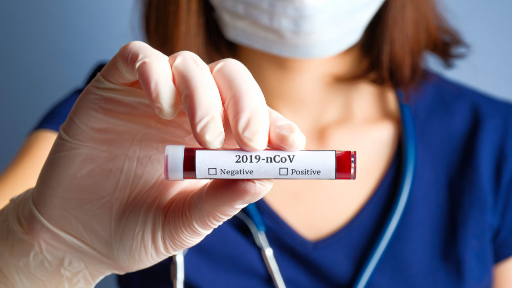 A masked nurce hold up a vial of blood labeled "2019-nCoV"