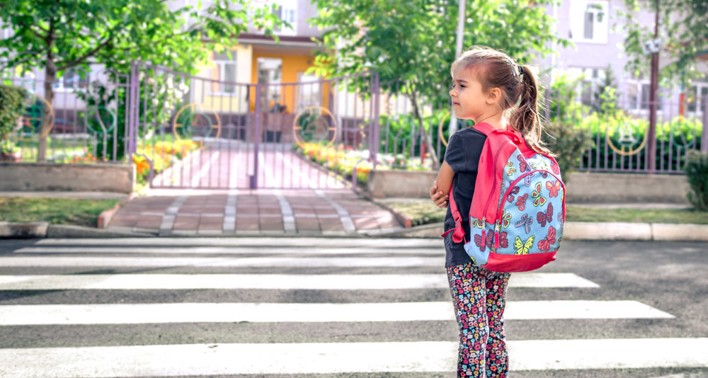 A young girl wearing a backpack stands by herself at a crosswalk while waiting to cross the street