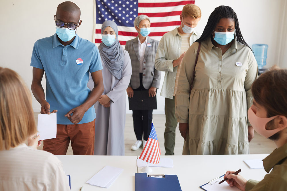 How To Get Involved In the 2020 Election Front view at multi-ethnic group of people standing in row and wearing masks at polling station on election day, focus on two African-American people registering for voting
