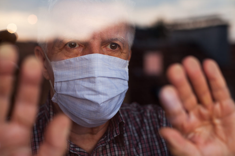 Elderly white man wearing face mask while looking out the window of a nursing home.