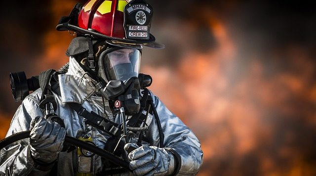 A fireman in a flame retardant suit stands with a large fire in the background