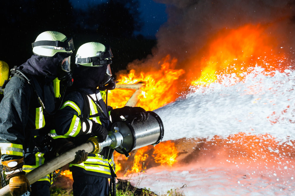 Two firefighters in full gear spray foam from a hose onto a raging fire