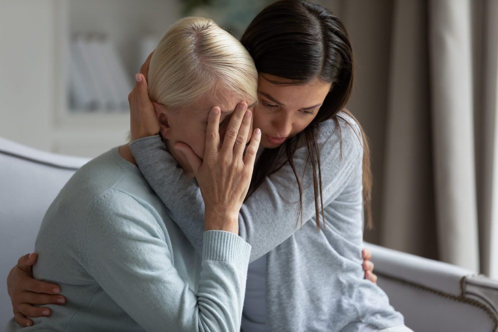 young lady embracing soothing crying depressed elder mother, sitting together at home.