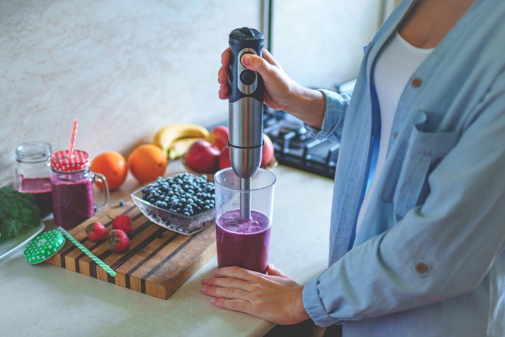 Woman cooking fresh blueberry smoothie with hand blender at kitchen at home. 