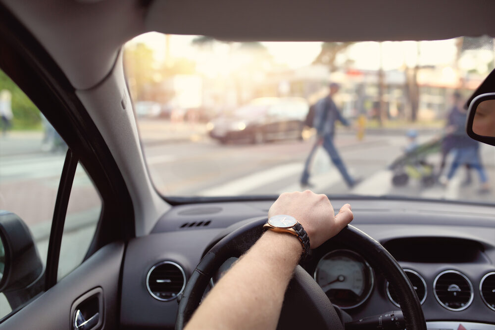 inside view of a car with a drivers hand on the steering wheel waiting for pedestrians to pass in crosswalk