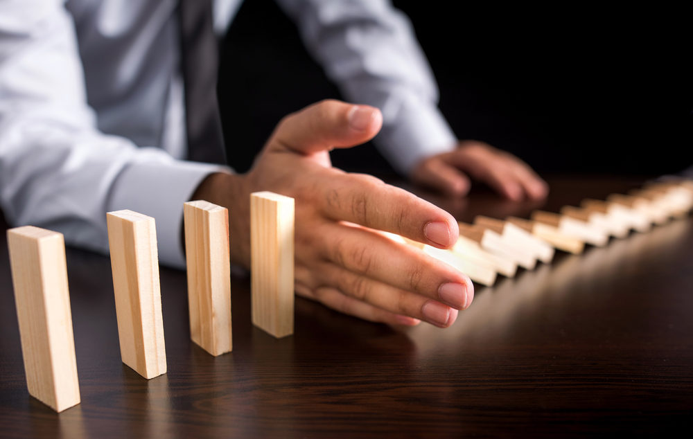 A male hand protects assets from the domino effect by stopping falling dominos on table