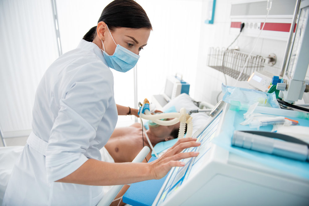 A mechanical ventilator being adjusted by a facemask-wearing nurse at a hospital