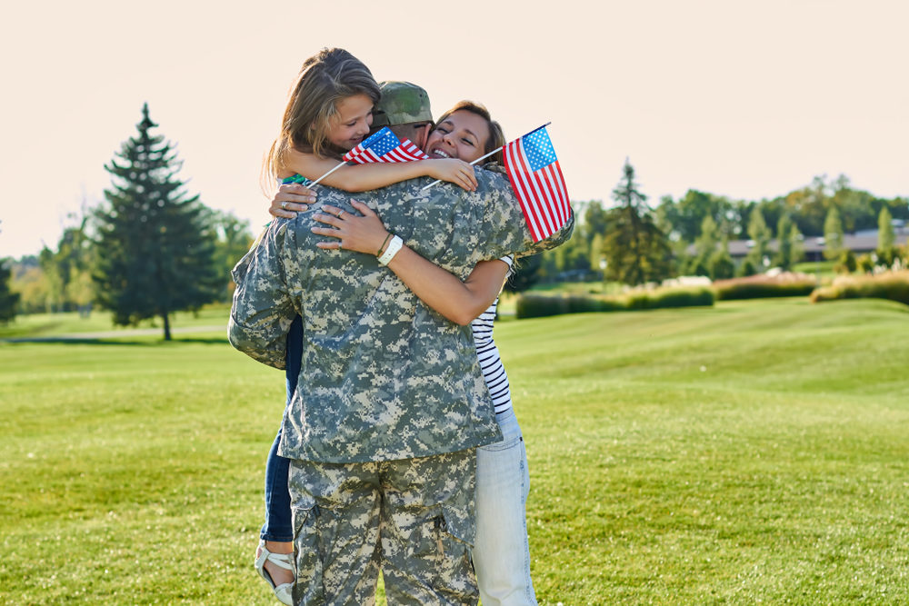 Military Vet Who Served at Camp Lejeune Sues Firefighting Foam Manufacturers Mother and daughter holding miniature flags embrace male US soldier in camouflage outside on a sunny day..