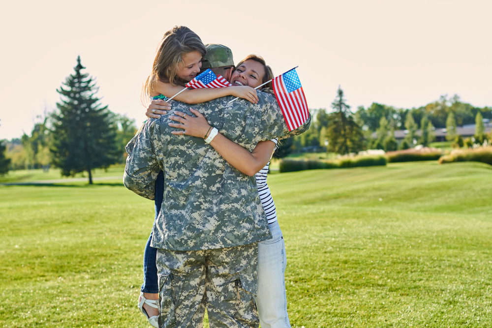 Mother and daughter holding miniature flags embrace male US soldier in camouflage outside on a sunny day..