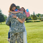Mother and daughter holding miniature flags embrace male US soldier in camouflage outside on a sunny day..