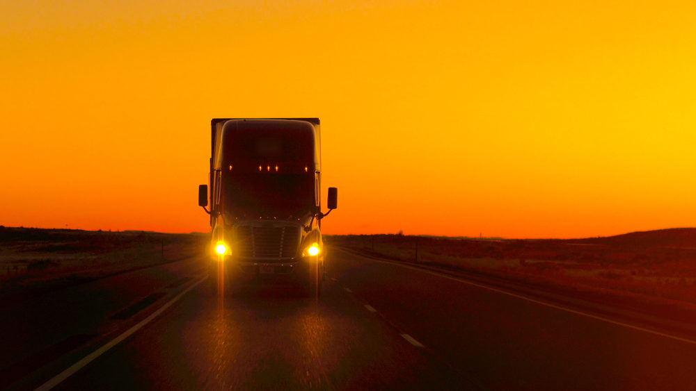 Semi truck driving and hauling goods on empty highway across the Great Plains in golden morning.