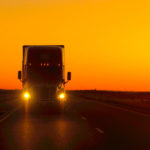 Semi truck driving and hauling goods on empty highway across the Great Plains in golden morning.