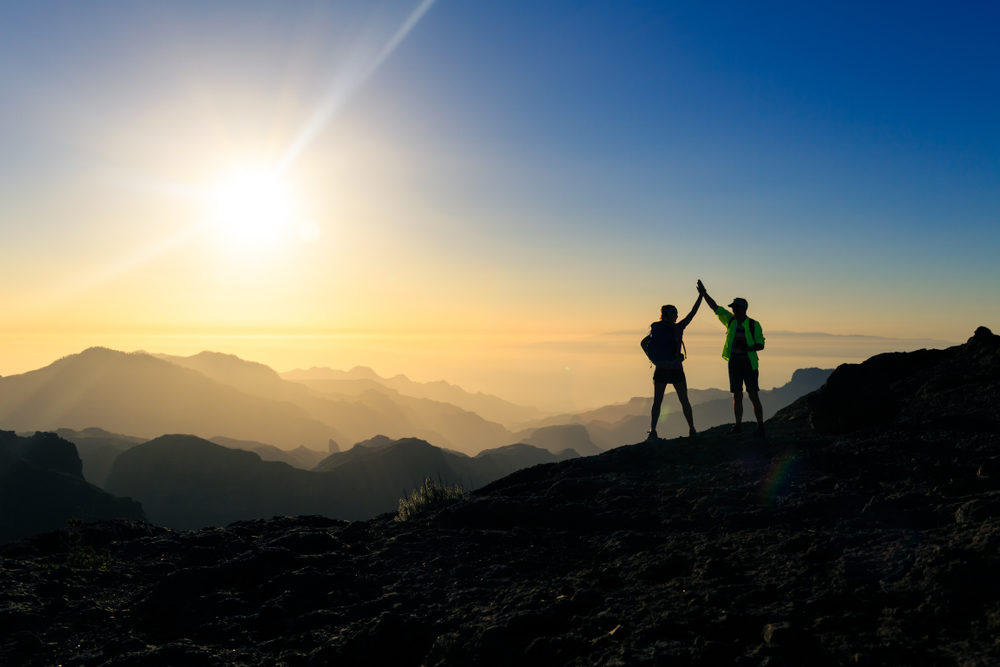 Couple hikers celebrating success in sunset mountains, accomplish with arms up outstretched