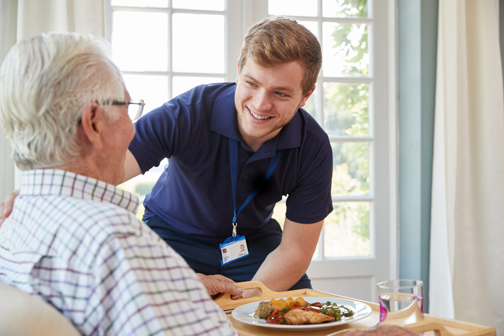 An elderly man is served dinner by a young male nurse at a home
