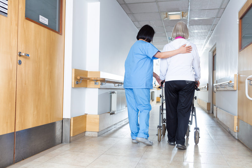A nurse helps an elderly woman with a walker walk down a hallway