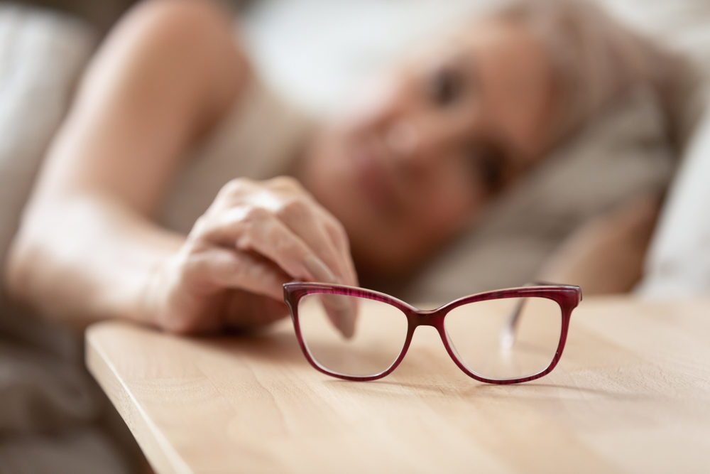 Older mature woman holding taking optical glasses from bedside table waking up in morning