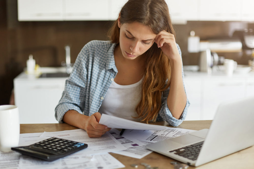 A young woman site at a desk with paperwork, a laptop, and a calculator