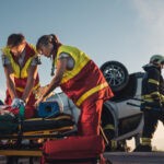 paramedics attending to a car accident victim on a stretcher with overturned vehicle behind them