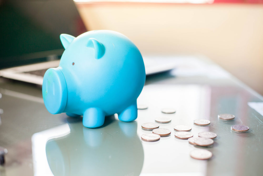 A cute blue piggy bank on a desk with coins beside it, laptop in the background