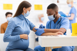 African American Doctor Giving Coronavirus Vaccine Injection Shot To Pregnant Woman, Wearing Protective Face Mask In Clinic Hospital. 