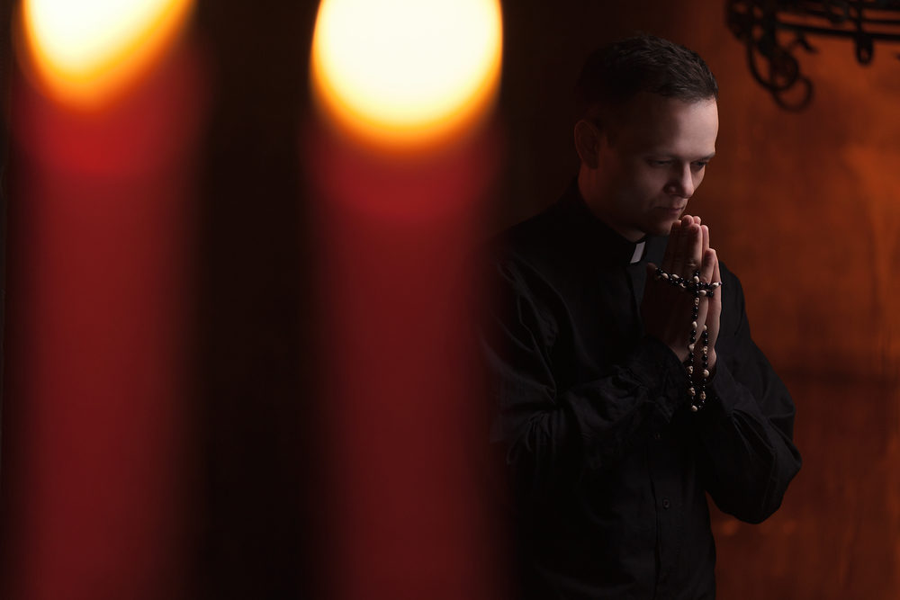 Diocese of Syracuse Commits to $100 Million to Settle Bankruptcy Claims Portrait of priest Next to the candles prays with his hands folded near the face.