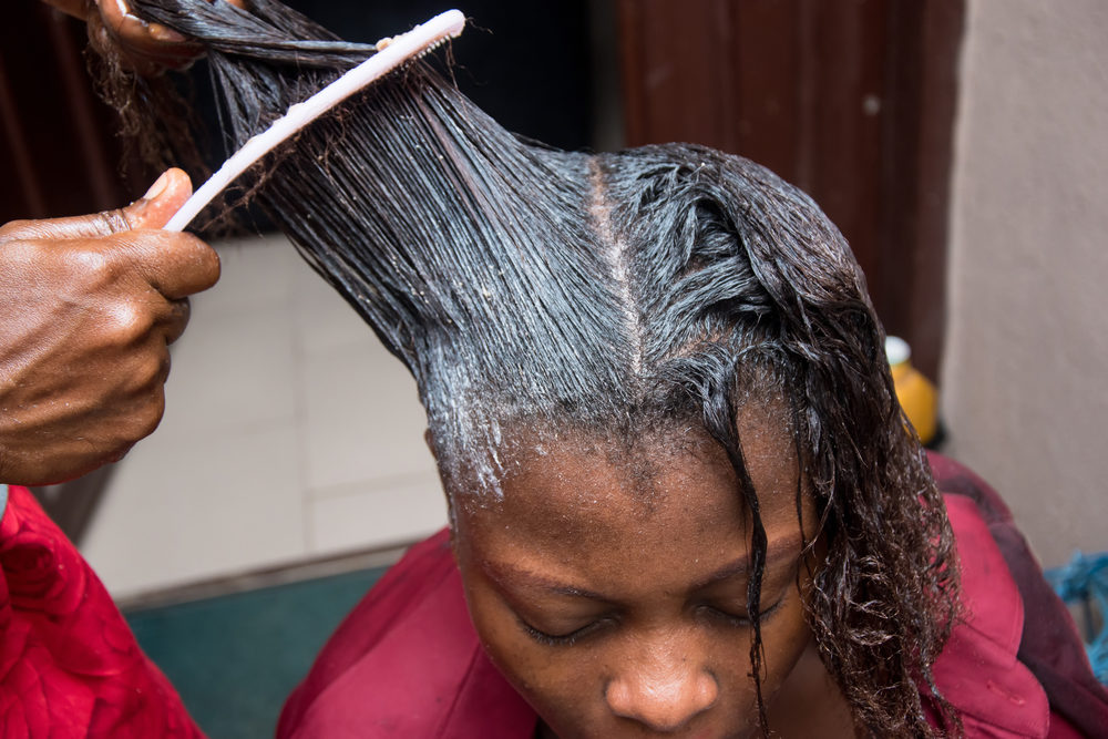 Hairdresser relaxing the hair on an African woman head and also using comb to stretch and apply the relaxer cream through the hair