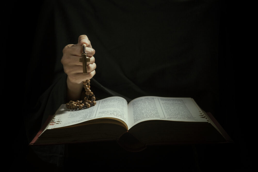 New Orleans Priest Charged with Sexual Assault from 1975 closeup of Hands holding rosary beads and cross while reading bible