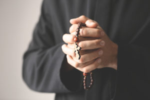 Young priest with rosary beads on light background