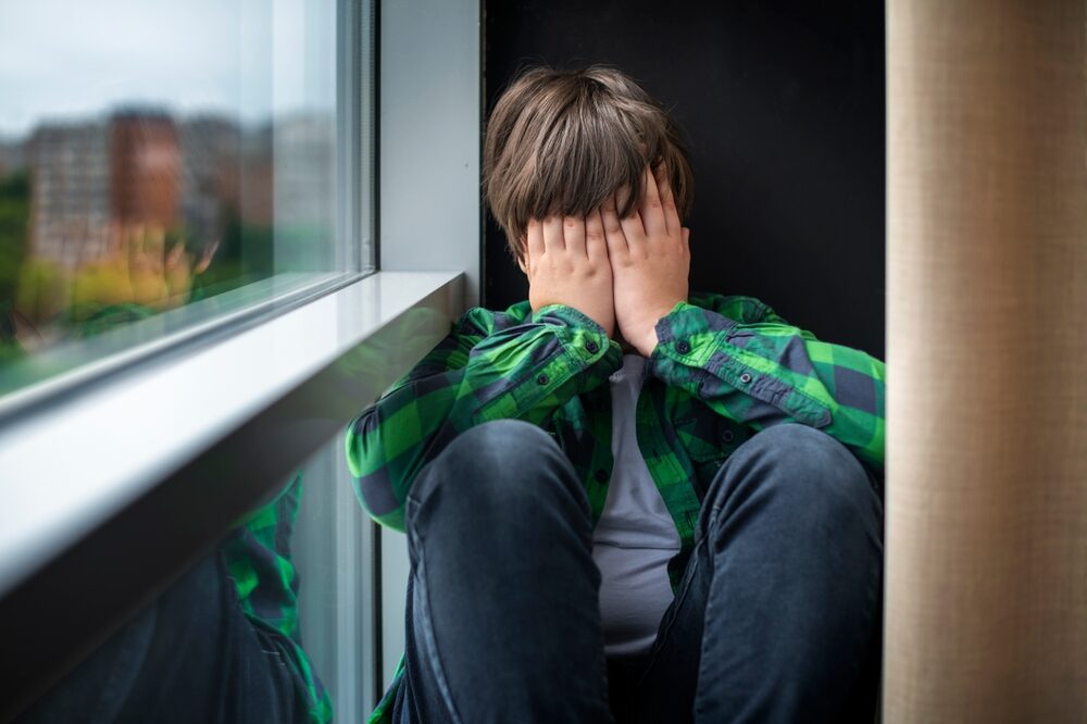 a sad teenage boy sitting on the windowsill at school with his hands over his face