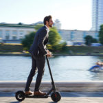 Businessman in a suit riding an e-scooter past a riverway